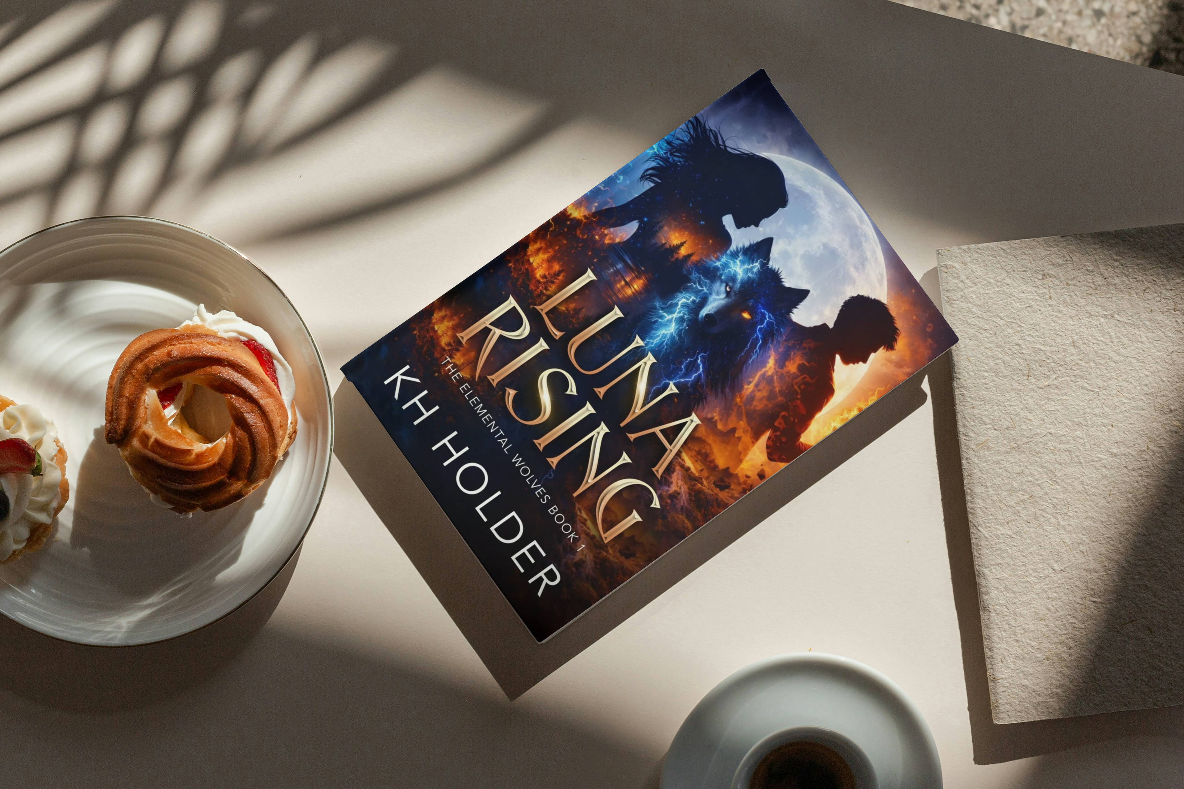 Book titled 'Luna Rising' by KH Holder on a table with a plate of dessert and a cup.